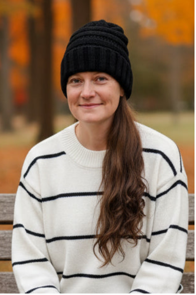 A woman wearing a black ribbed knit beanie and a cream sweater with black horizontal stripes, sitting on a wooden bench outdoors in a park with autumn trees in the background.