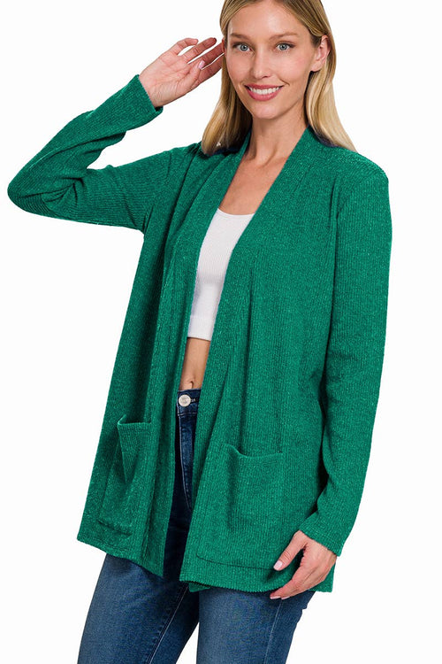 A woman wearing a heather green open-front cardigan with large front patch pockets, styled over a white crop top and blue jeans, against a plain white background.