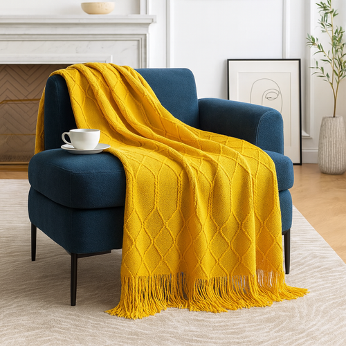 A bright yellow knit throw blanket with fringe is draped over a deep blue armchair in a styled living room setting with a fireplace, rug, and framed art.