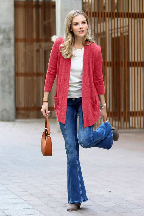 A woman wearing a coral pink open-front cardigan, blue flare jeans, and a white top walks outdoors while holding a brown leather handbag.