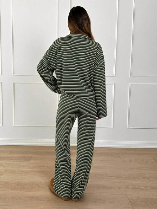 A woman viewed from behind wearing a matching two-piece pajama set with thin horizontal stripes in olive green and white, standing on a light wood floor against a white paneled wall.