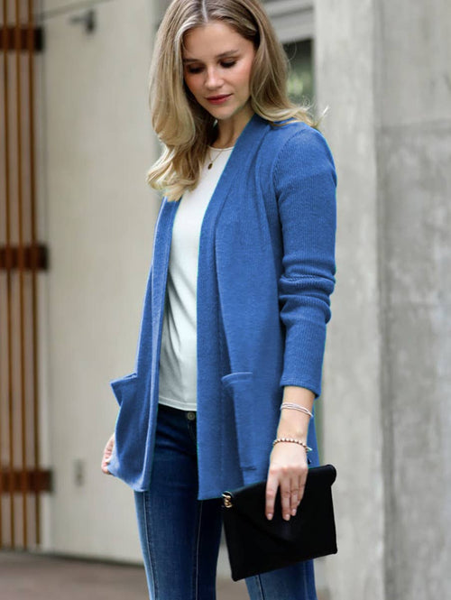 A woman wearing a blue open-front cardigan over a white top and blue jeans, holding a black clutch bag in an outdoor urban setting.