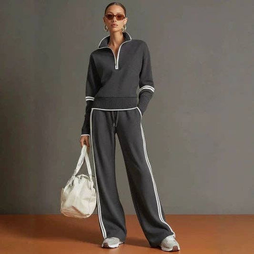 A model wearing a dark gray two-piece tracksuit with white contrast stitching, featuring a half-zip mock-neck sweater and matching wide-leg pants, holding a white tote bag against a gray studio background.