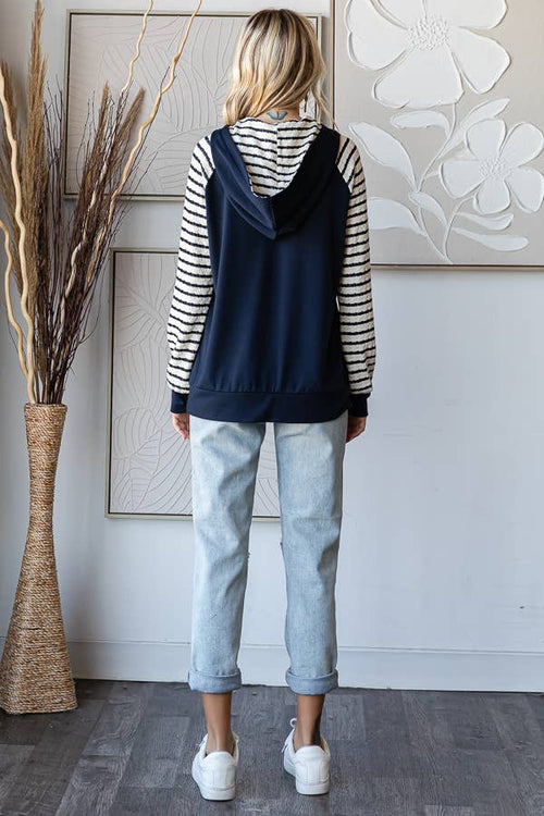 A woman is shown from behind wearing a navy blue hooded sweatshirt layered over a black and white striped long-sleeve shirt, paired with light-wash blue jeans and white sneakers, standing in a modern room with a decorative vase and wall art.