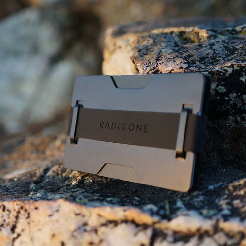 A slim metallic card holder or wallet with the brand name RADIX ONE debossed on the front, resting on a textured natural rock outdoors.