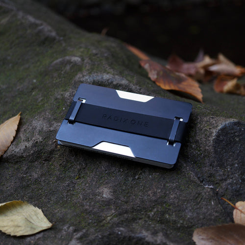 A slim black and silver metallic card holder or wallet with the brand name 'RADIX ONE' embossed on the front, resting on a mossy gray rock outdoors with scattered dry leaves.