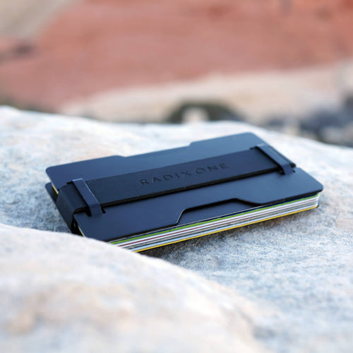 A slim black metal wallet card holder with the brand name RADIXONE embossed on the top, holding several cards, resting on a light gray stone with a blurred outdoor background.