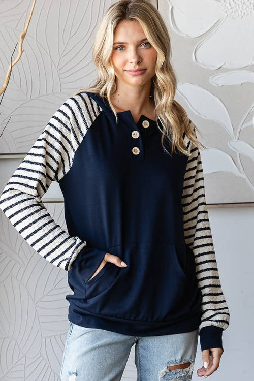 A woman modeling a long-sleeve henley sweatshirt with navy blue body and black-and-white striped sleeves, paired with light-wash jeans, standing against a light gray wall with leafy wall art.