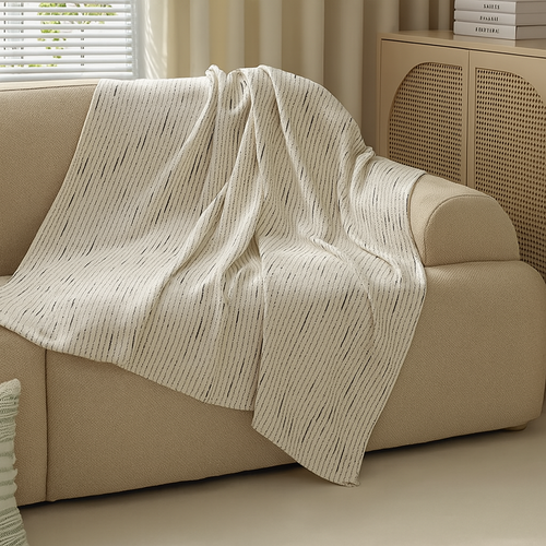 A cream-colored throw blanket with thin vertical black stripes is draped over a beige fabric sofa in a softly lit living room setting.