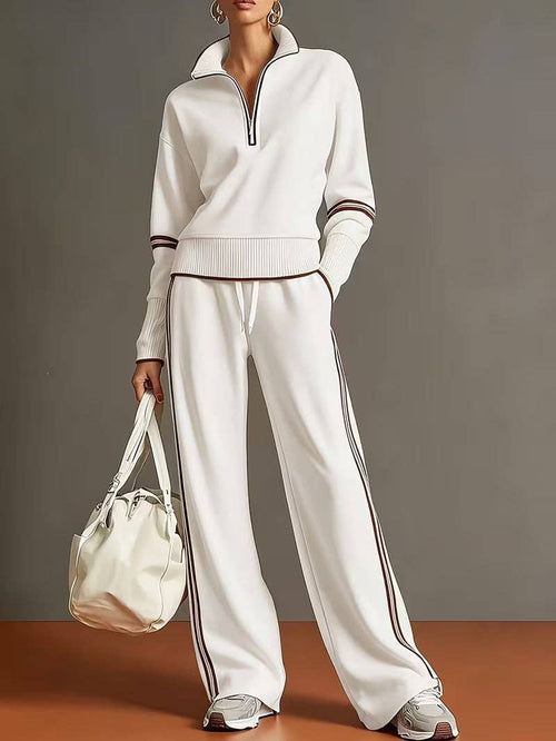 A model wearing a matching white half-zip pullover and wide-leg sweatpants with brown stripe accents, holding a white handbag against a gray studio background.