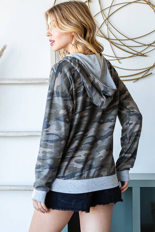 A woman wearing a gray and black camouflage hoodie with a heather gray ribbed hem and cuffs, paired with dark denim shorts, shown from a side-back angle against a white wall with decorative wire art.