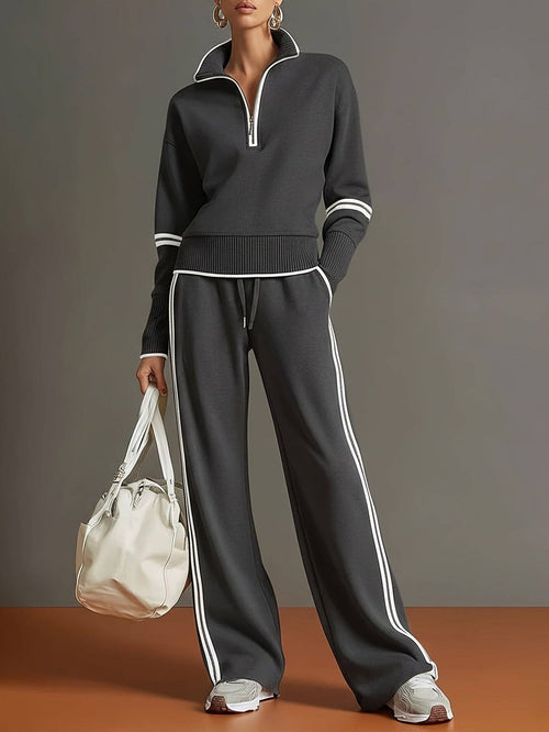 A model wearing a dark gray ribbed knit tracksuit with white stripe accents on the sleeves, hem, and pants, holding a white tote bag against a gray and terracotta studio background.