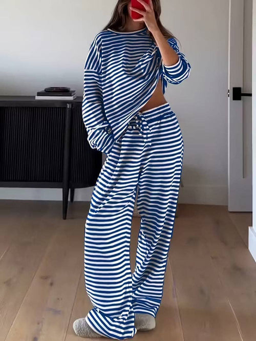 A woman wearing a matching blue and white striped lounge set consisting of a cropped long-sleeve top and wide-leg pants, standing in a room with light wood floors.