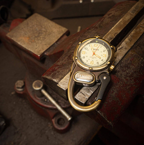 A vintage-style wristwatch with a gold-tone case and a black carabiner-style strap attachment, resting on a worn red and steel bench vise.