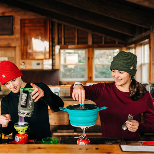 Two people cooking outdoors using portable stoves in a wooden cabin kitchen.