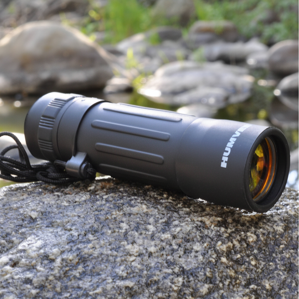 A black HUMMING single-prism binocular sight resting on a wet rock outdoors, with a blurred natural background of rocks and water.