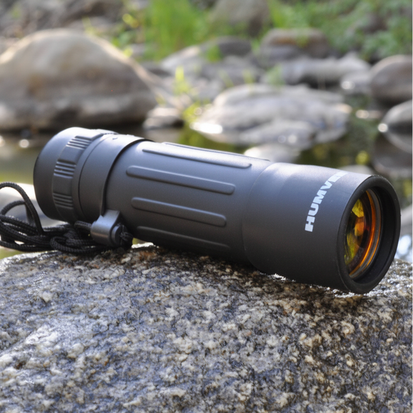 A black HUMMING single-prism binocular sight resting on a wet rock outdoors, with a blurred natural background of rocks and water.