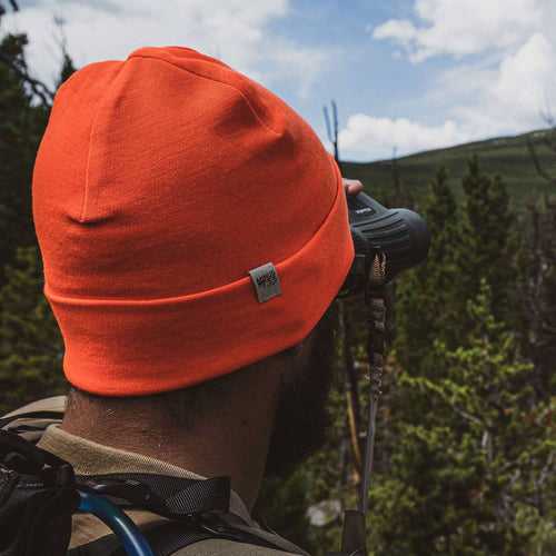 A man outdoors wearing a bright orange beanie with a small gray tag that reads 'MP35', seen from behind as he looks through binoculars. Trees and hills are visible in the background.