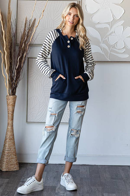 A woman modeling a navy blue raglan sweatshirt with black and white striped sleeves, paired with distressed light-wash jeans and white sneakers, standing in a modern room setting.
