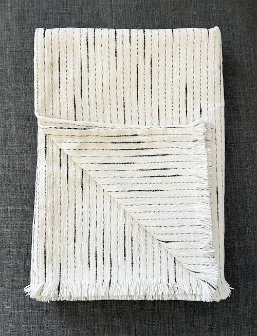 A folded off-white woven throw blanket with black vertical stripe patterns and fringed edges, displayed on a dark gray textured fabric background.