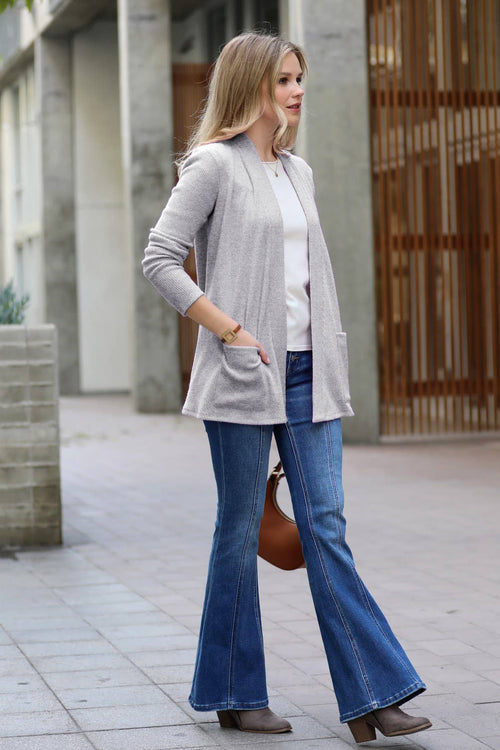 A woman stands outdoors wearing a light gray open-front cardigan over a white top, blue flared jeans, and brown ankle boots, with a brown handbag at her side.