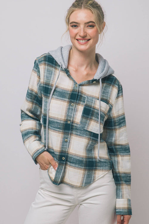 A woman wearing a hooded plaid flannel shirt with a gray hoodie lining and white pants, standing against a light gray studio background.