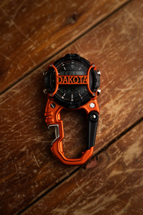 An orange and black digital compass with a carabiner attachment, featuring the brand name 'DAKOTA' on the dial, resting on a rustic wooden surface.