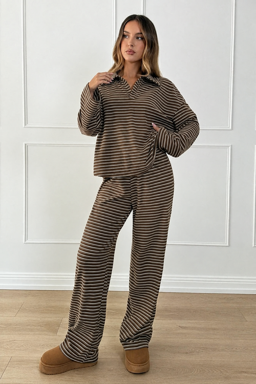 A woman modeling a matching two-piece lounge set with a brown and beige striped long-sleeve collared top and wide-leg pants, paired with tan platform slippers, standing in front of a white paneled wall.