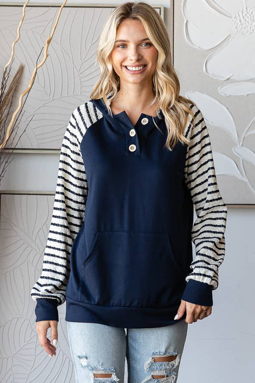 A woman wearing a navy blue pullover hoodie with black and white striped raglan sleeves, paired with light-wash distressed jeans, standing in front of a neutral-toned wall with decorative dried branches.