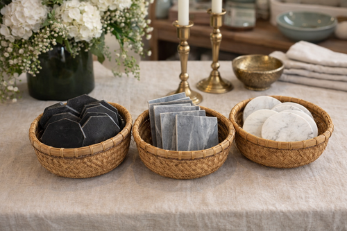 Three woven wicker baskets arranged on a linen tablecloth, holding black soap bars, gray stone tiles, and white marble coasters, with a vase of white flowers and brass candlesticks in the background.