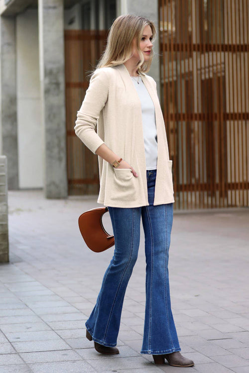 A woman stands outdoors wearing a cream open-front cardigan over a white top, blue flared jeans, and brown ankle boots, holding a brown handbag.