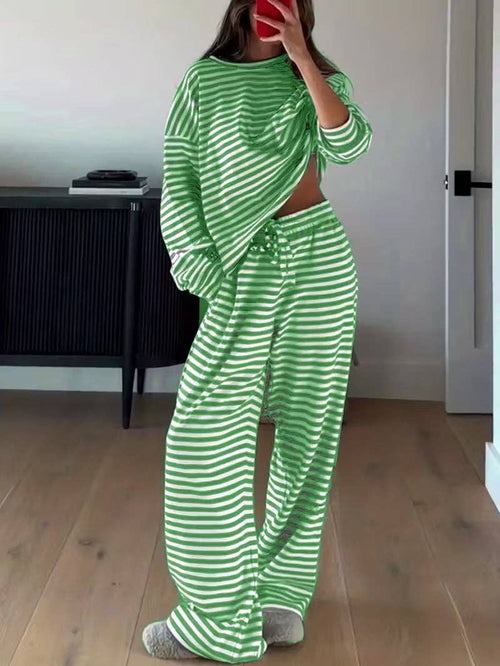 A woman modeling a matching two-piece lounge set with green and white horizontal stripes, consisting of a cropped long-sleeve top and wide-leg pants, standing in a room with light wood floors.