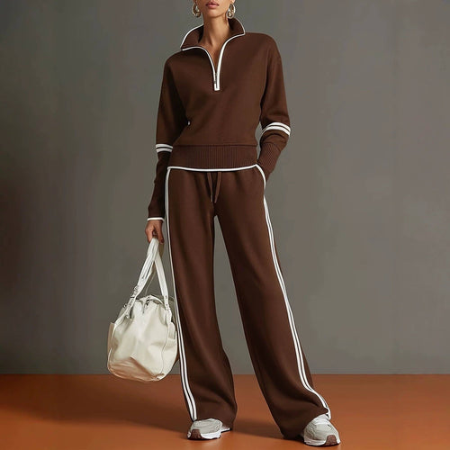 A model wears a matching brown knit two-piece tracksuit with white contrast trim, featuring a half-zip long-sleeve top and wide-leg pants with side stripes. She holds a white bucket bag against a neutral studio backdrop.