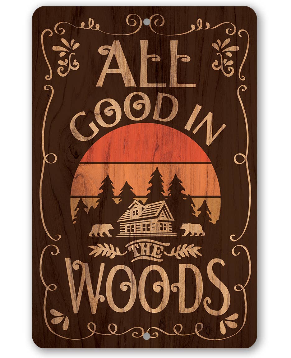 A rectangular wooden sign with rounded corners featuring the phrase 'ALL GOOD IN THE WOODS' in light tan lettering over a dark brown woodgrain background. The center shows a stylized sunset with an orange and tan sun, pine trees, a cabin, and bears.