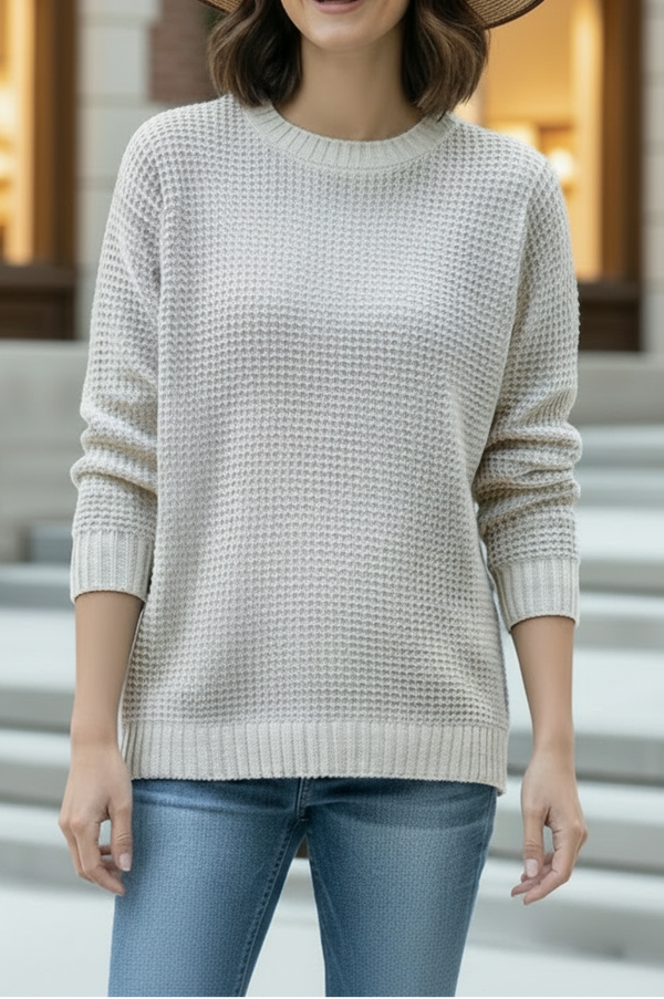 A woman wearing a cream waffle-knit crewneck sweater, blue jeans, a beige wide-brim straw hat, and sunglasses, standing in front of a blurred brick building with steps.