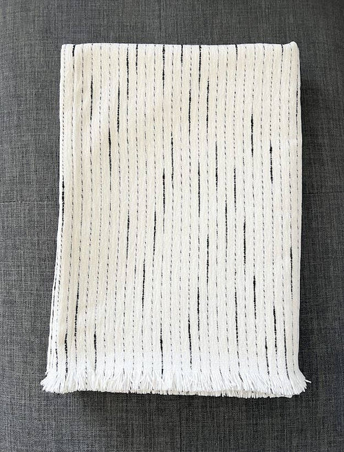 A folded off-white woven throw blanket with black vertical dash patterns and fringed edges, displayed on a dark gray textured fabric background.