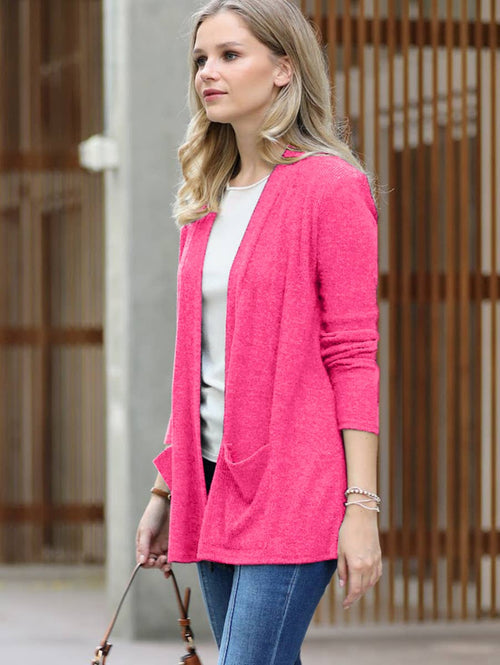 A woman wearing a bright pink open-front cardigan over a white top and blue jeans, standing in front of a modern wooden slatted wall.