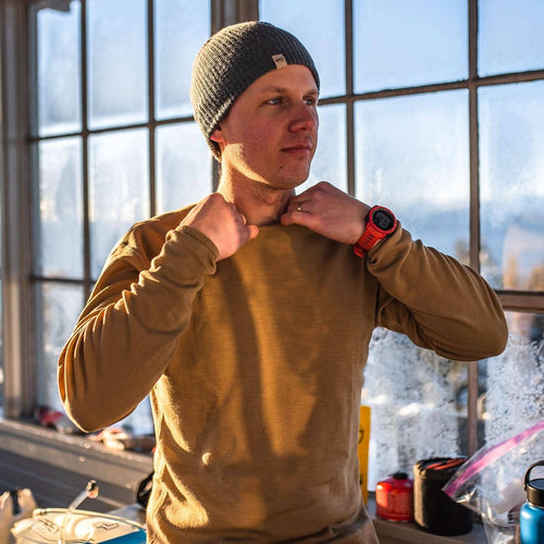 A man wearing a dark gray knit beanie and a tan long-sleeve performance shirt stands in front of a large window with snow visible outside. He is adjusting the collar of the shirt and wearing a red smartwatch.