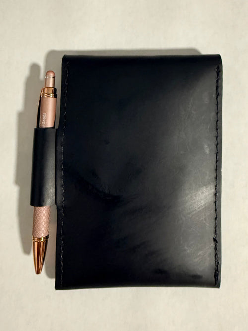 A black leather notebook cover or wallet with a pen loop holding a rose gold pen, photographed from above on a light off-white surface.