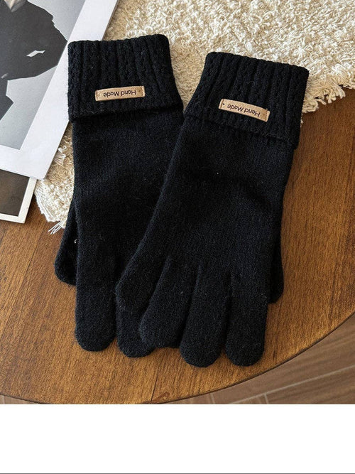 A pair of black knit touchscreen-compatible gloves with a small tan brand label on the cuff, displayed on a wooden surface next to a textured rug.