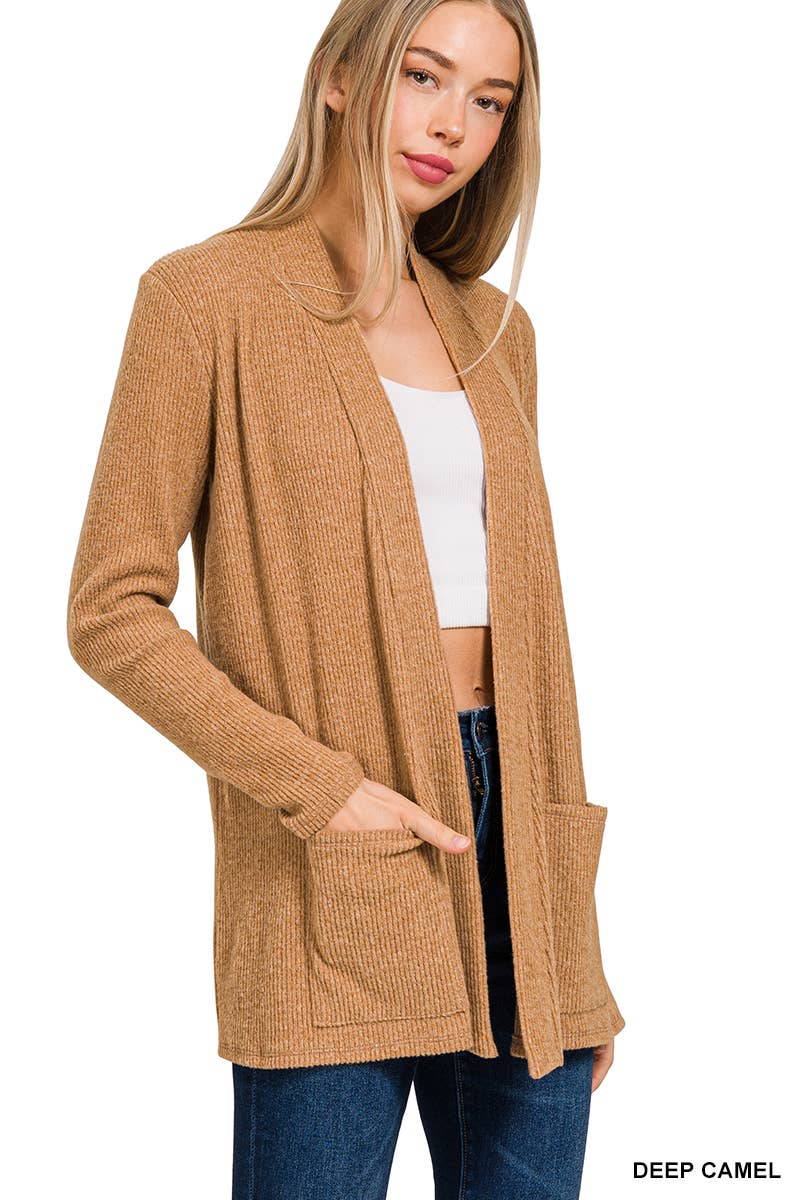 A woman modeling a long open-front ribbed cardigan in a warm camel tan color, worn over a white crop top and blue jeans against a plain white background.