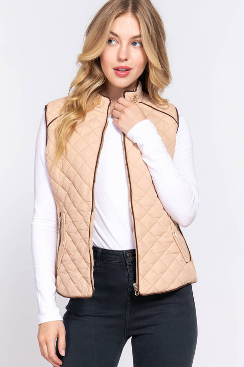A woman wearing a beige quilted zip-front vest over a white long-sleeve top and black jeans, photographed against a light gray studio background.