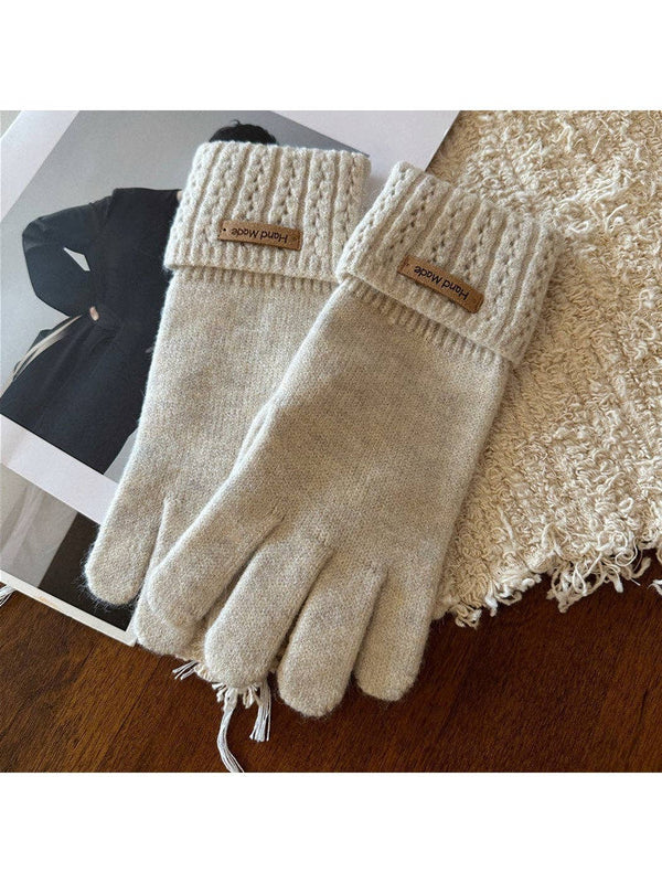 A pair of cream-colored knit touchscreen gloves with ribbed cuffs and small brown brand patches, displayed on a wooden surface next to a textured blanket.