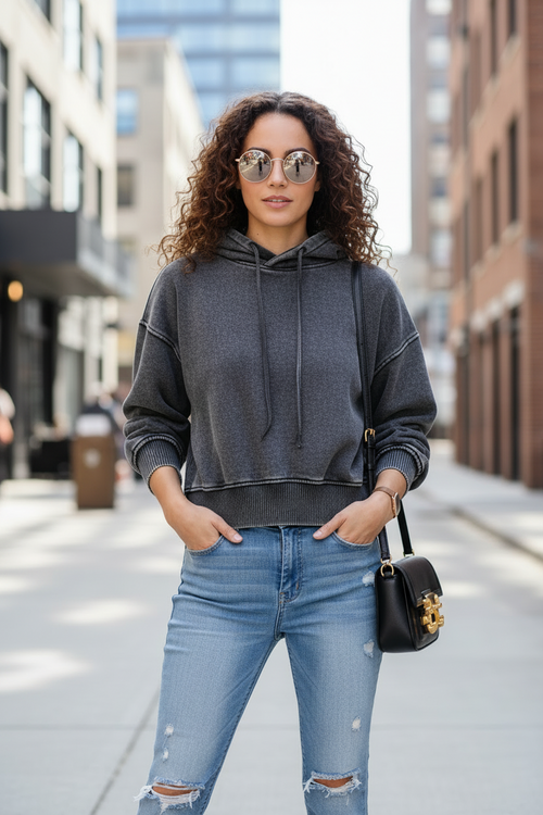 A woman wearing a dark gray cropped hoodie and light blue ripped jeans stands on a city street, holding a black crossbody bag with a gold clasp.