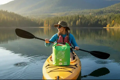 Woman kayaking on calm lake with forested mountains, wearing hat and life vest, green dry bag on yellow kayak