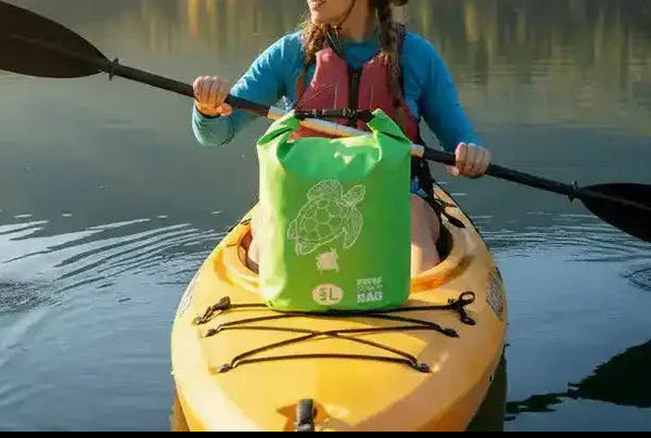 Woman kayaking on calm lake with forested mountains, wearing hat and life vest, green dry bag on yellow kayak
