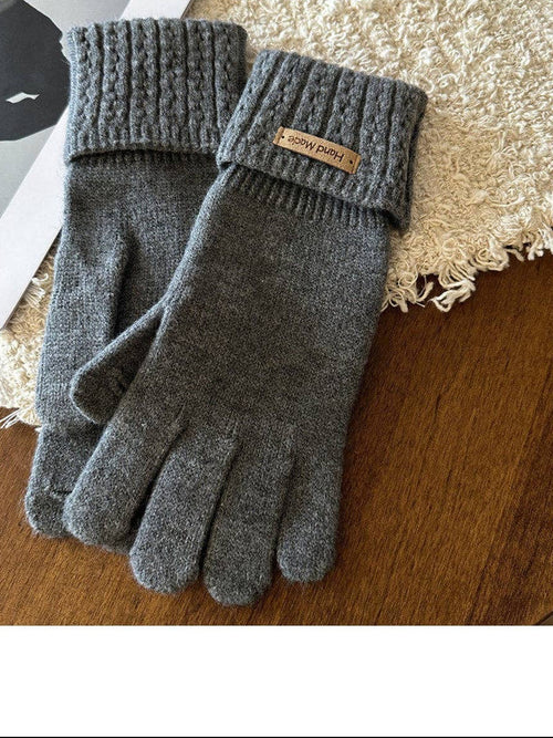 A pair of dark gray knit fingerless gloves with ribbed cuffs, displayed on a wooden surface next to a cream shag rug and a magazine page. A small tan leather brand tag on the cuff reads 'SUNNY DAYS'.