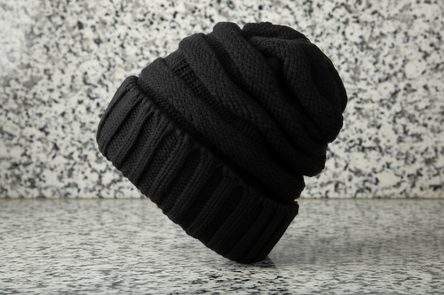 A black chunky knit beanie hat resting on a light wood surface against a neutral light gray background.