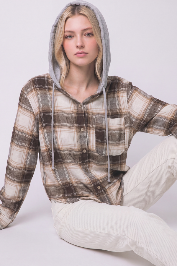 A woman wearing a brown and beige plaid hooded button-up shirt with a gray-lined hood, paired with off-white pants, seated against a light gray studio background.