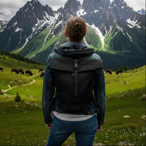 A woman viewed from behind wearing a black backpack with a flap closure, standing in a lush green mountain meadow with grazing cows and dramatic snow-capped peaks in the background.